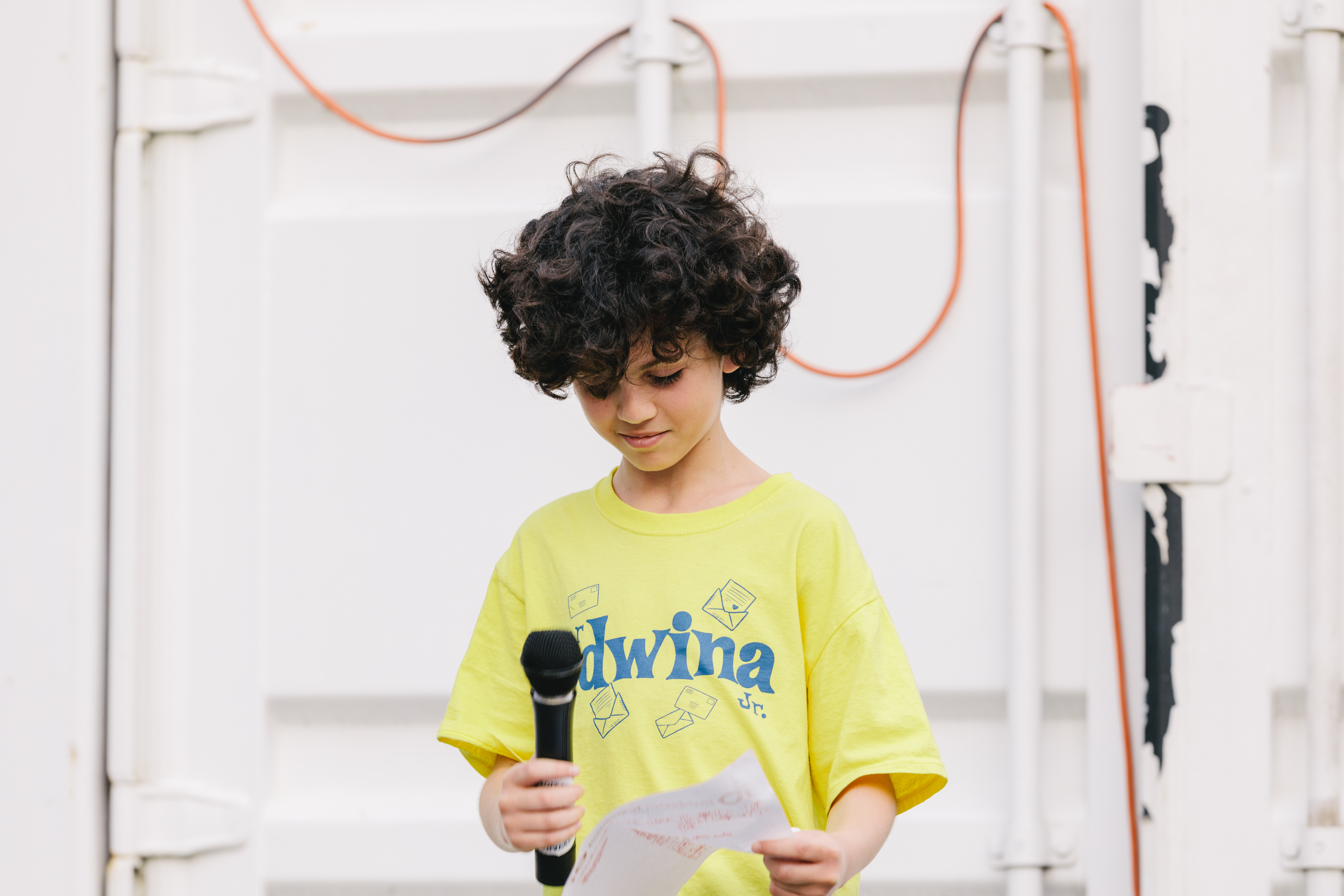 Child with brown curly brown hair reads at an outdoor open mic in front of a shipping container.