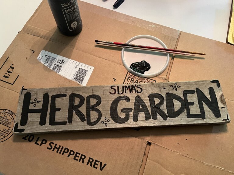 Wooden sign reading "SUMA'S HERB GARDEN" in black painted letters lies on top of a cardboard box. A paintbrush and black paint are beside it.