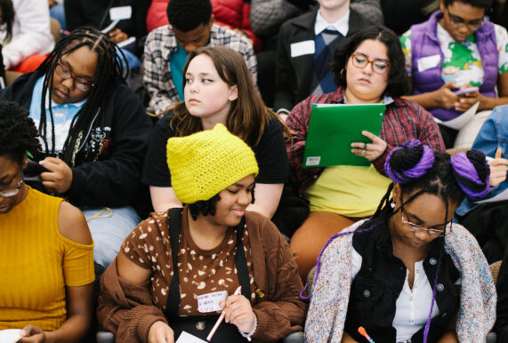 Group of teens sits in an auditorium talking and writing at the 2023 Detroit Youth Poetry Con