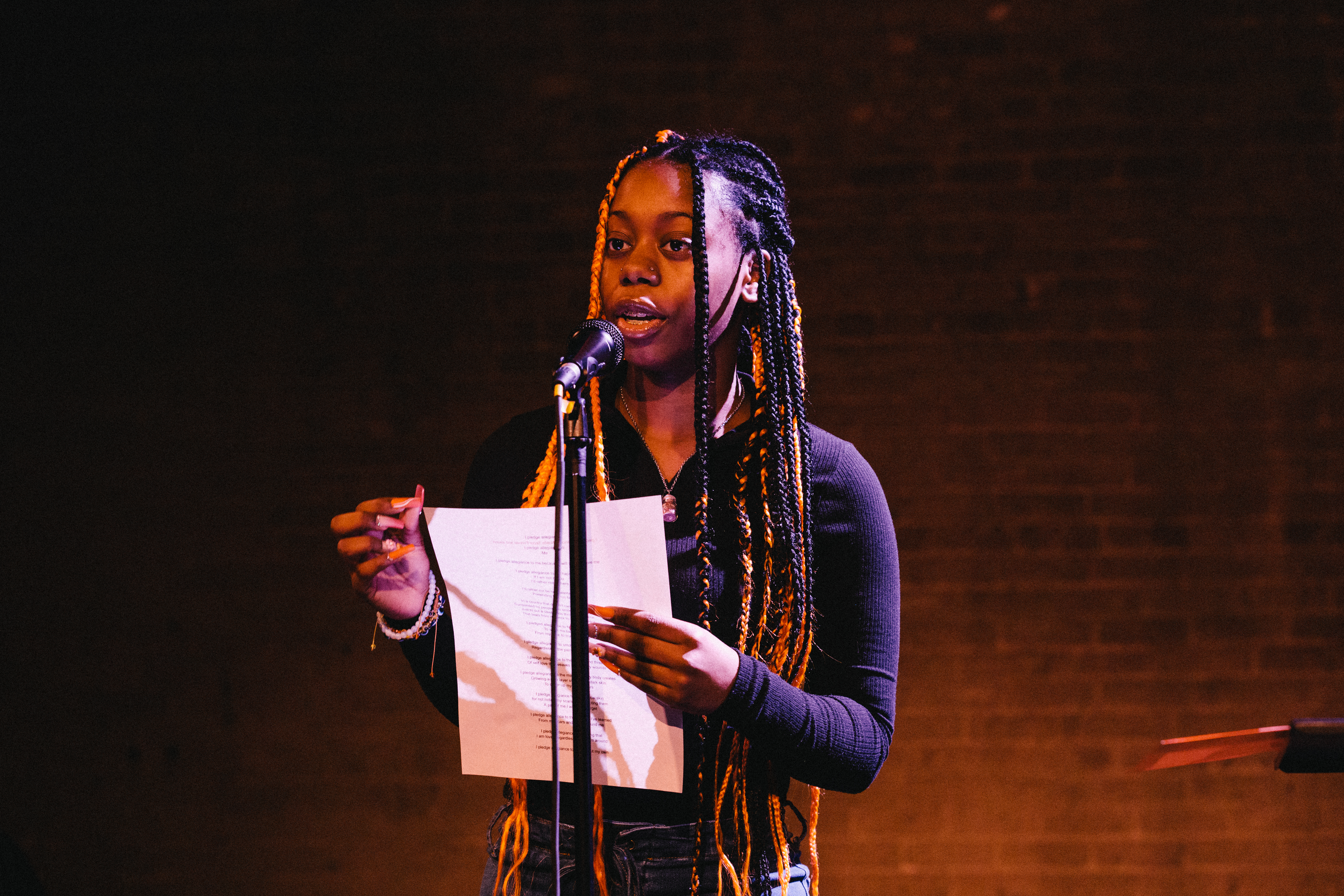 InsideOut student poet performs at the public showcase in 2023. She has long braids and holds a poem on paper at a microphone against a dark background.