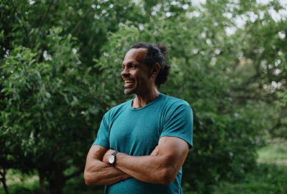 Ross Gay stands in a green t-shirt with his arms crossed, smiling, outside against a group of trees.