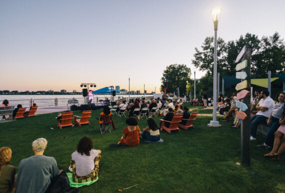 A crowd of people sit in orange Adirondack chairs facing the Detroit river at sunset.