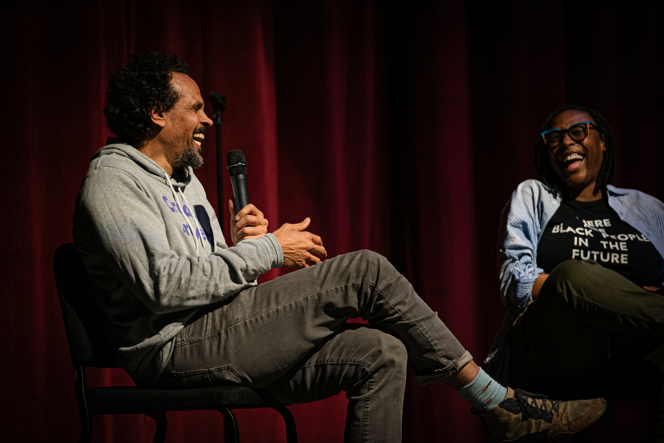 Ross Gay and Nandi Comer are seated and laughing onstage in a dark auditorium.