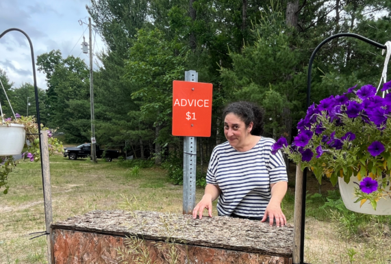 Suma Karaman Rosen stands at a wooden stand at the side of the road with a red sign behind her that reads "Advice $1."