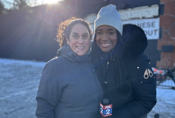 Executive Director Suma Karaman Rosen and Fox 2 Detroit Reporter Liz Lewin are wearing parkas and backlit by a rising sun on a cold winter day, against a wall of brick painted in a dark color.