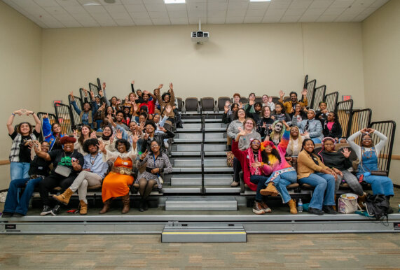 Teens at Detroit Youth Poetry Con sit on risers in a large room smiling and waving their hands excitedly!